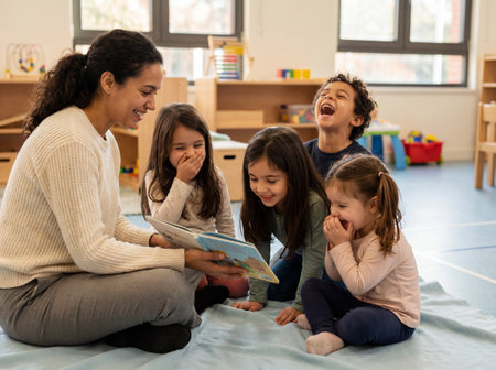A cheerful female teacher sits on the floor reading a funny storybook to a diverse group of four happy children who are laughing together in a bright modern kindergarten classroom.の素材