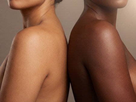 Two diverse women stand back to back to highlight the texture and contrast between their smooth bare shoulders and their natural glowing skin complexions.の素材