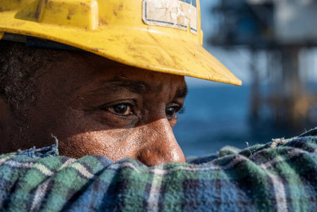 A close up detail of an african american male worker wearing a dirty yellow hard hat and plaid shirt resting on his arms while looking pensive with a blurred ocean rig in the background.の素材