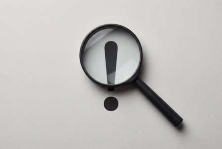 A black magnifying glass lies on a white paper background focusing on a large black exclamation point to symbolize discovery, attention, warning, significant findings and search results.の素材