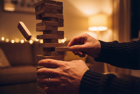 A person's hands carefully removing a block from a precarious tower of wooden blocks in a game of skill, with a warm and cozy atmosphere in the backgroundの素材