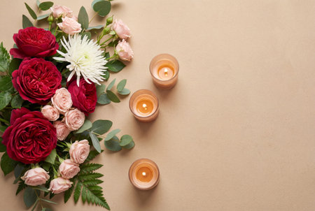 Top view of a bouquet containing deep red roses, pink spray roses, white chrysanthemums and eucalyptus leaves next to three lit candles on a beige surface with ample copy space.の素材