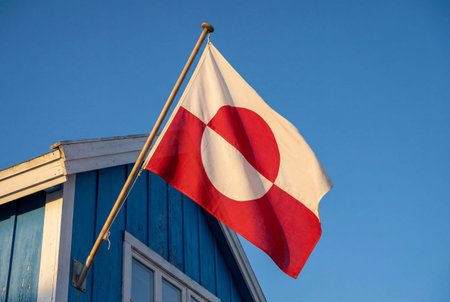 The red and white national flag of greenland hangs from a wooden pole attached to the side of a traditional bright blue wooden house against a clear blue sky during a sunny day.の素材