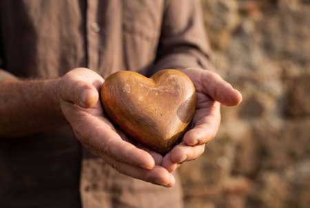 A man wearing a brown shirt gently holds a smooth carved wooden heart in his cupped hands against a blurred background symbolizing love care and charity.の素材
