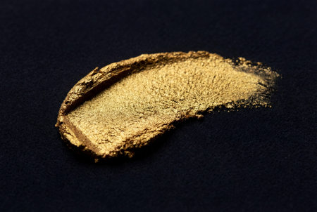 A single stroke of metallic gold makeup showing rich texture and sparkling grain against a dark velvety black background surface for luxury beauty concepts.の素材