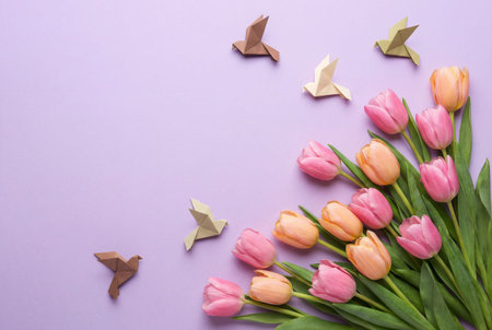 A creative spring composition featuring fresh pink and orange tulips arranged with handmade origami paper doves on a smooth pastel purple background with copy space for text.の素材