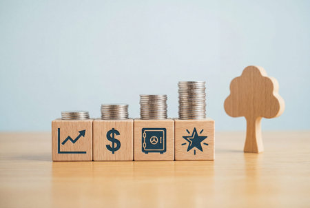 Light oak blocks with financial icons of rising graph, dollar sign, vault, and star arranged beside growing stacks of coins and tree shape on desk for wealth accumulation conceptの素材