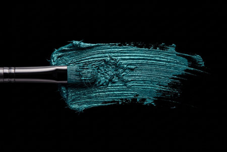 A cosmetic brush drags a thick layer of metallic teal cream across a black background creating a textured swatch with shimmering emerald pigments and fibrous details.の素材