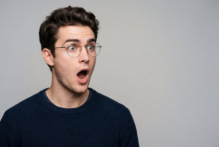 Young adult man with flawless glowing skin wearing round eyeglasses shows surprised open mouth reaction displaying shocked yet intrigued facial expressionの素材