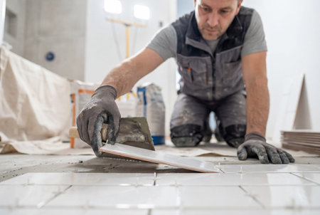A construction worker is laying white ceramic tiles on a floor using spacers. home improvement, flooring installation, and renovation service by a professional tilerの素材