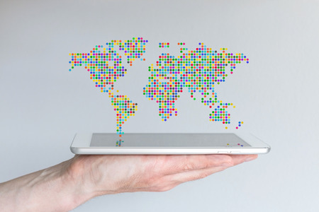 World map floating above a modern smart phone or tablet. Hand holding mobile device in front of gray background. Reflections on screen. Concept of mobile computing and global communication.の写真素材