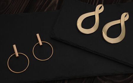 Golden girl accessories on dark background. Modern gold earrings pairs on dark stone background