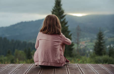 Little blonde girl sitting on terrace and looking at sunset behind the mountainsの写真素材
