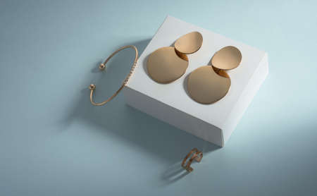 Top view of golden earrings and bracelet on white box on blue background
