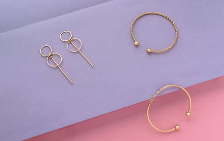 Top view of modern gold bracelets and earrings on purple paper on pink background