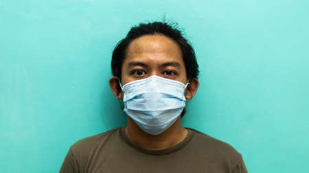 A portrait of a young Malay man wearing 3 layer surgical face mask on isolated blue background....