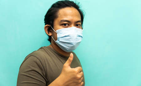 A portrait of a young Malay man wearing 3 layer surgical face mask, showing thumbs up on isolated blue background. Protection step to avoid spreading viruses and diseases.の写真素材