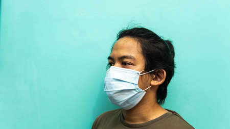 The side view portrait of a young Malay man wearing 3 layer surgical face mask on isolated blue background. Protection step to avoid spreading viruses and diseases. Medical concept.の写真素材
