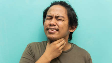 A portrait of a Malay man holding his neck, sore throat with painful face reaction on isolated blue background. Medical concept. Selective focused.の写真素材