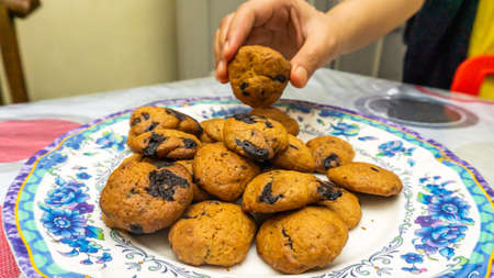 Woman hand taking one piece of cookies with chocolate chips from the stacks of cookies on the plate at the table. Homemade dessertの写真素材