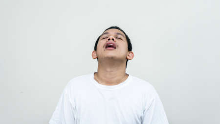 A portrait of tired Asian Malay man with white t-shirt closing eyes sighed tired and annoyed on isolated white backgrounds. Bad mood with heavy work.の写真素材