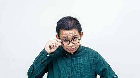 Confused face on an Asian Malay man with green shirt concentrate looking at the camera holding pair of spectacles on isolated white background. Checking out the subject in front of him.の写真素材