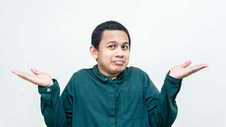 A portrait of clueless Asian Malay man funny smiling in green shirt, shrugging with hands in different sides, smirking anf being unaware what is happening on isolated white background.の写真素材