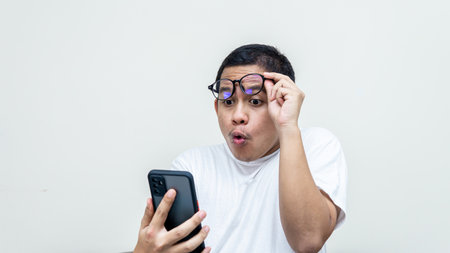 Wow and surprised expression of young Asian Malay man with spectacles in white t-shirt by looking something he saw on the smartphone screen on isolated white background.の写真素材