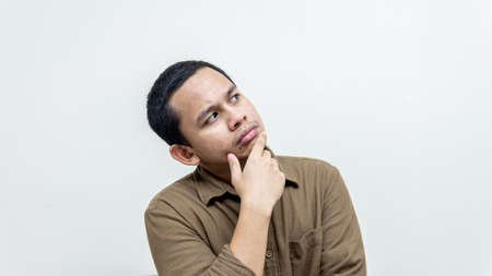 Confused and thinking face expression of young Asian Malay man with hand on the chin looking on empty space with casual brown shirt with isolated white background. Thinking idea concept.の写真素材