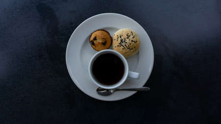 Top view photo, a white cup of americano black coffee, cup cake and cereal bread on white plate on black backgroundの写真素材