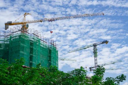 Upward view of precast building and large tall Tower Crane moving machine in construction work, scaffolding cover by green net protection on each story, green trees on foreground under white fluffy clouds blue skyの写真素材