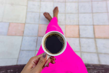 A white cup of black coffee in a tan skin woman's hand wearing fresh pink dress, sitting on a wooden chair on grey and brown tiles floorの写真素材