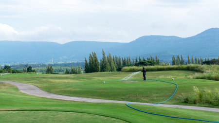 Landscape of green grass Lawn yard, golf  course with a worker watering the lawn land, mountain in background under cloudy skyの写真素材