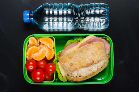 Sandwich, small tomatoes, tangerines in plastic lunch box and bottle of water on black chalkboard.