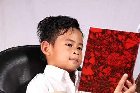 Smart Asian boy reading book while sitting on chairの写真素材