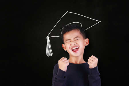 Success concept, portrait of happy young Asian student boy showing enthusiastic winning gesture, shout with joy of victory, over blackboard with graduation hat drawn above his headの写真素材