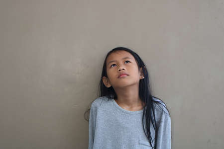 Little Asian girl standing against grey wall, looking to the sky and thinking somethingの写真素材