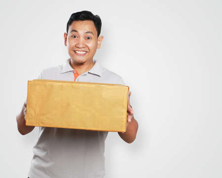 Photo image portrait of a cute handsome young Asian courier man smiling while giving gift package box in delivery dutyの写真素材
