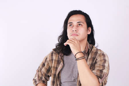 Photo image portrait of a cute young Asian male student with long hair, looking up seriously, touching his chin and thinkingの写真素材