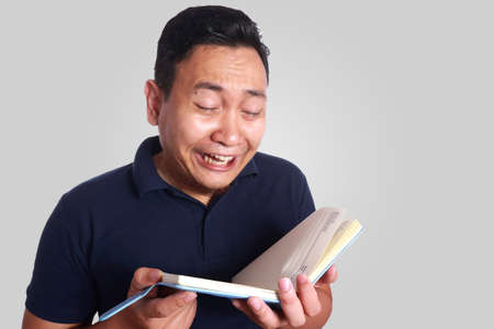 Photo image of cute funny Asian man crying while reading book on his handの写真素材