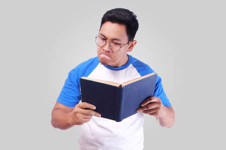 Photo image portrait of young funny Asian man reading book with angry gesture over grey background