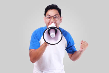Photo image portrait of funny young Asian man shouting with megaphone, mad yelling screaming crazy supporting motivating conceptの写真素材