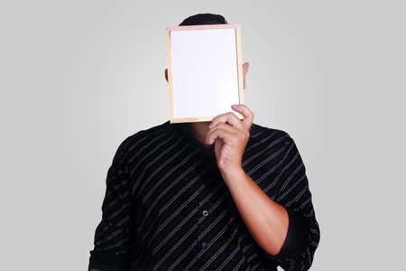 Portrait of a man wearing black shirt, holding and hiding covering his face with small empty whiteboard, copy space maskの写真素材