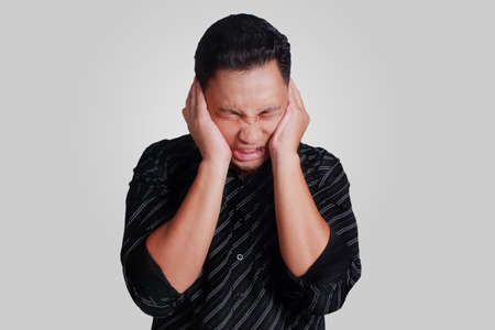 Portrait of young attractive Asian male closing his ears, disturbed to hear noises 