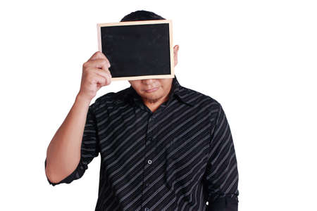 Portrait of young attractive Asian man wearing black shirt, holding and showing small empty copy space blackboard covering his face, isolated on whiteの写真素材