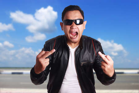 Portrait of young attractive angry Asian men wearing black leather jacket and sunglasses showing middle finger, over cloudy blue sky outdoor scenery backgroundの写真素材