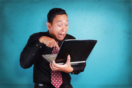 Portrait of young Asian businessman typing on laptop, funny excited expression, over blue wall backgroundの写真素材