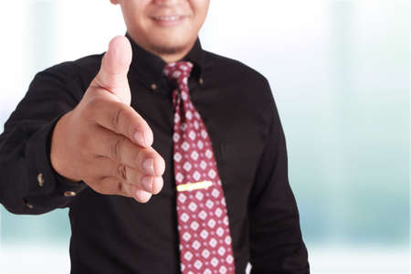 Close portrait of attractive young Asian businessman offering handshake, focus on fingers, over...