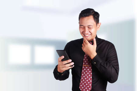 Portrait of young Asian businessman smiling happy to seed good news on his smartphone, over blurred...