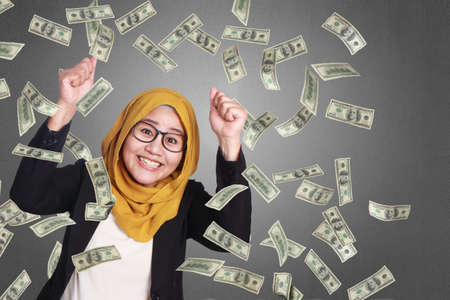 Portrait of success beautiful muslim businesswoman wearing hijab and eyeglasses winning victory gesture under rain of moneyの写真素材